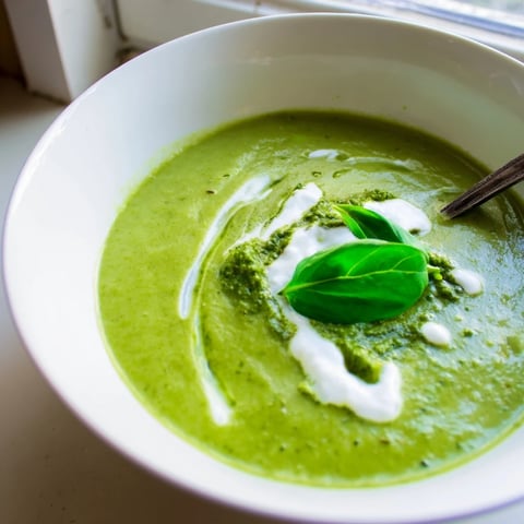 A vibrant bowl of Courgette, Pea and Pesto Soup garnished with a dollop of crème fraîche and fresh basil leaves.