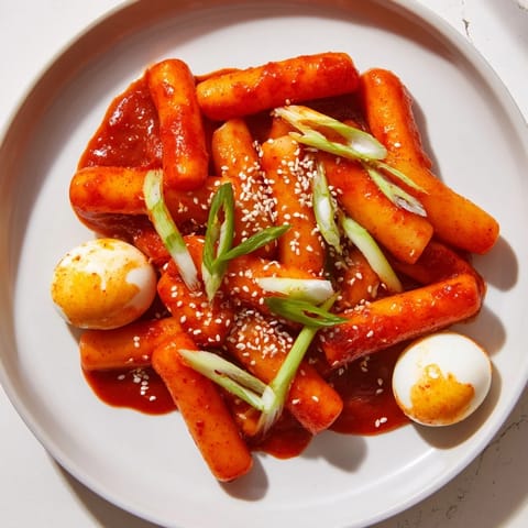 A close-up of chewy rice cakes coated in a glossy, sweet, and savory red sauce, with sliced onions and green onions for texture.  