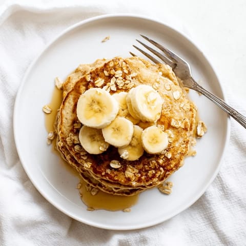 Golden-brown Banana Oat Pancakes simmering on a griddle with bubbling batter and a spatula ready to flip.  